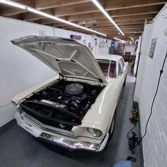 White classic Ford Mustang with bonnet open showing engine bay in a workshop garage