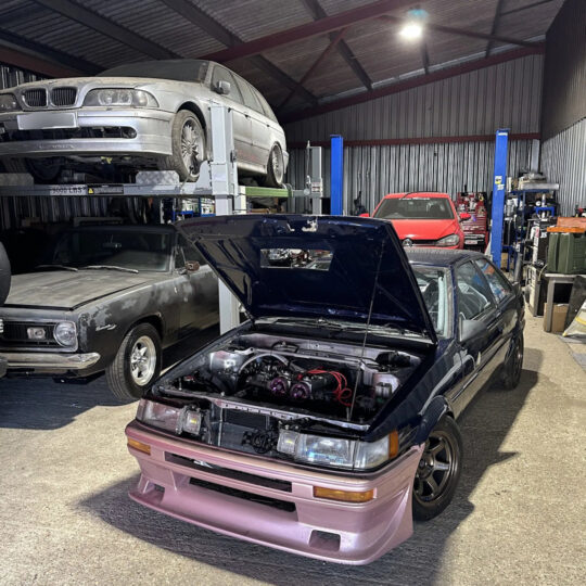 Modified classic car with bonnet open showing engine bay inside a garage workshop