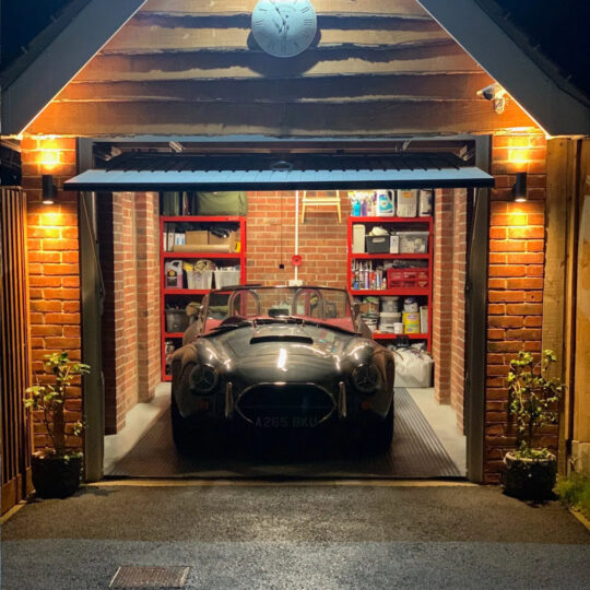 Warmly lit car cave at night with classic Cobra sports car visible through open garage door